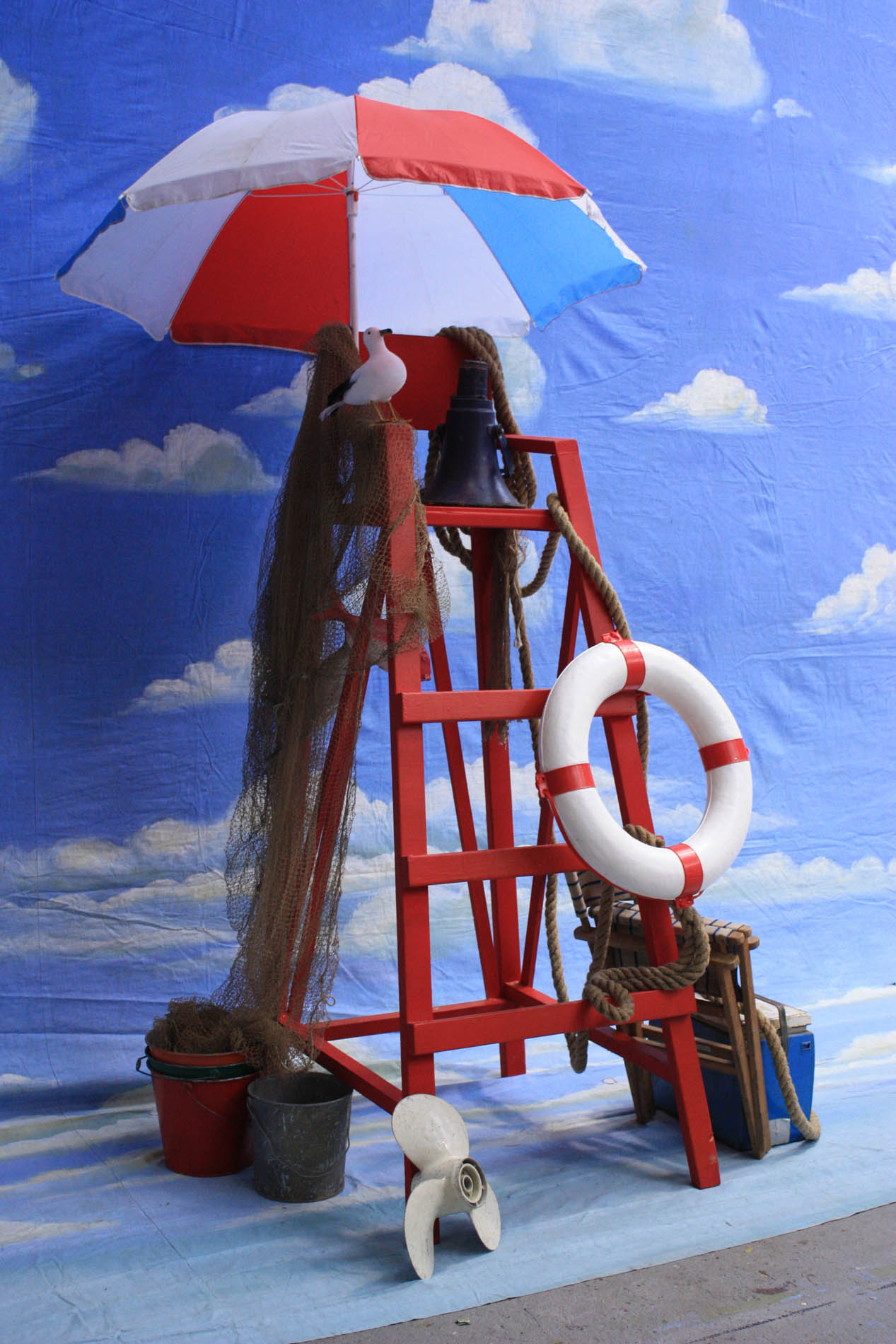 Red lifeguard tower draped in fishing net with a striped umbrella, life ring, buckets and an outboard propeller against a painted sky