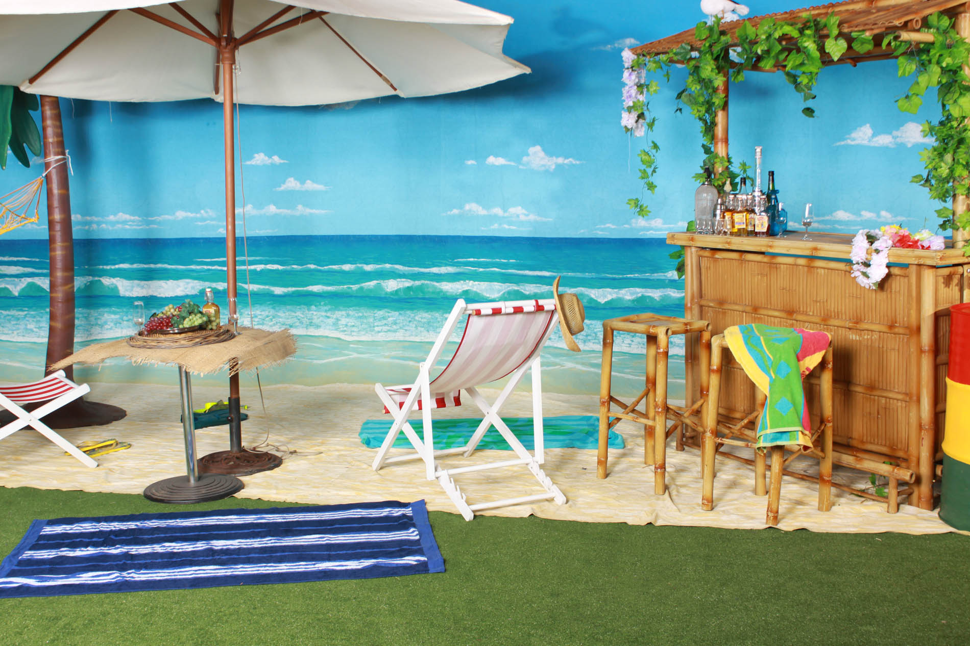 Beach bar set with a timber bar, bar stools, market umbrella, deck chair and a painted tropical beach backdrop