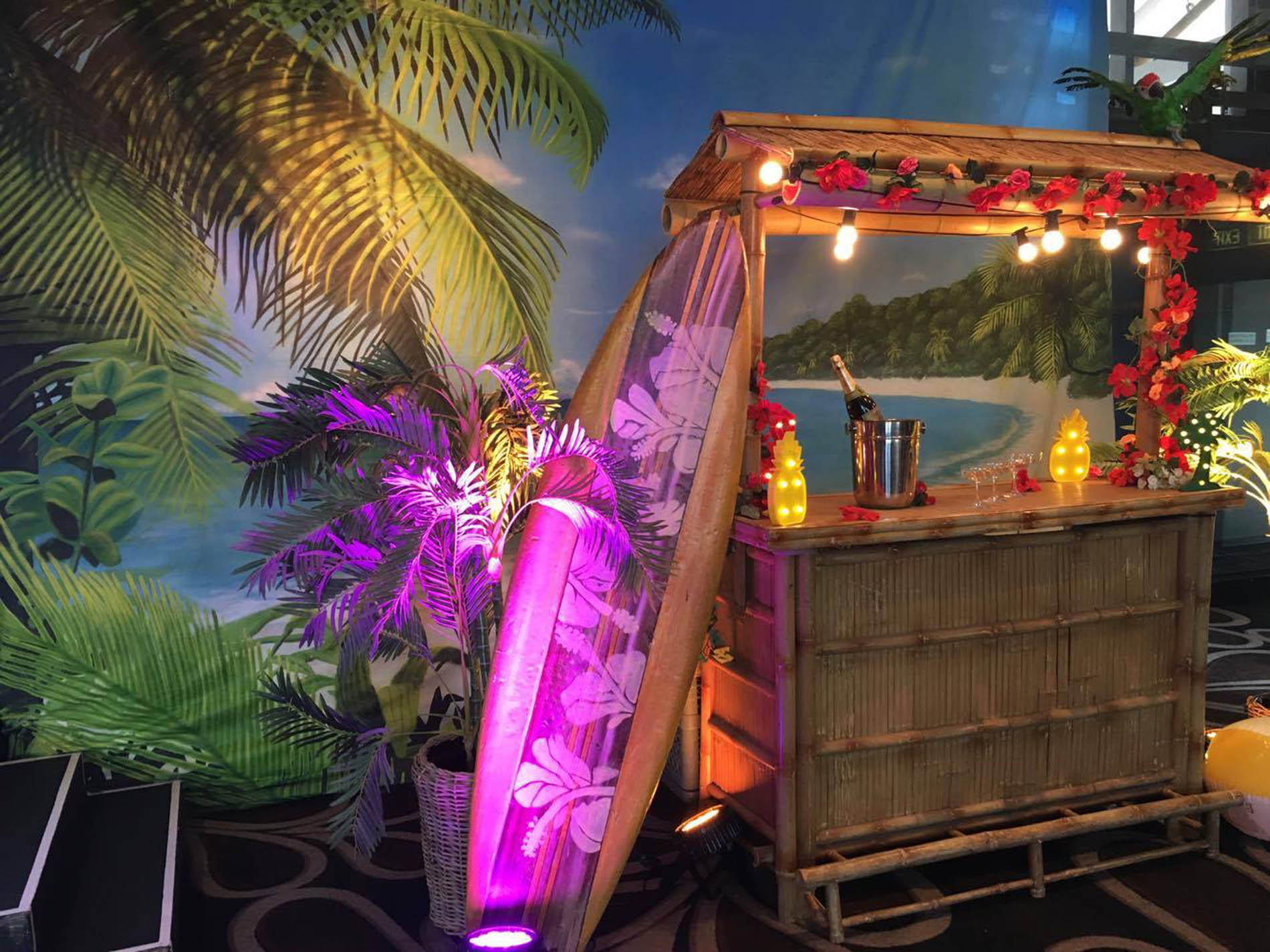 Bamboo tiki bar with festoon lights and tropical flowers, a palm-printed surfboard lit in purple, and lush palms