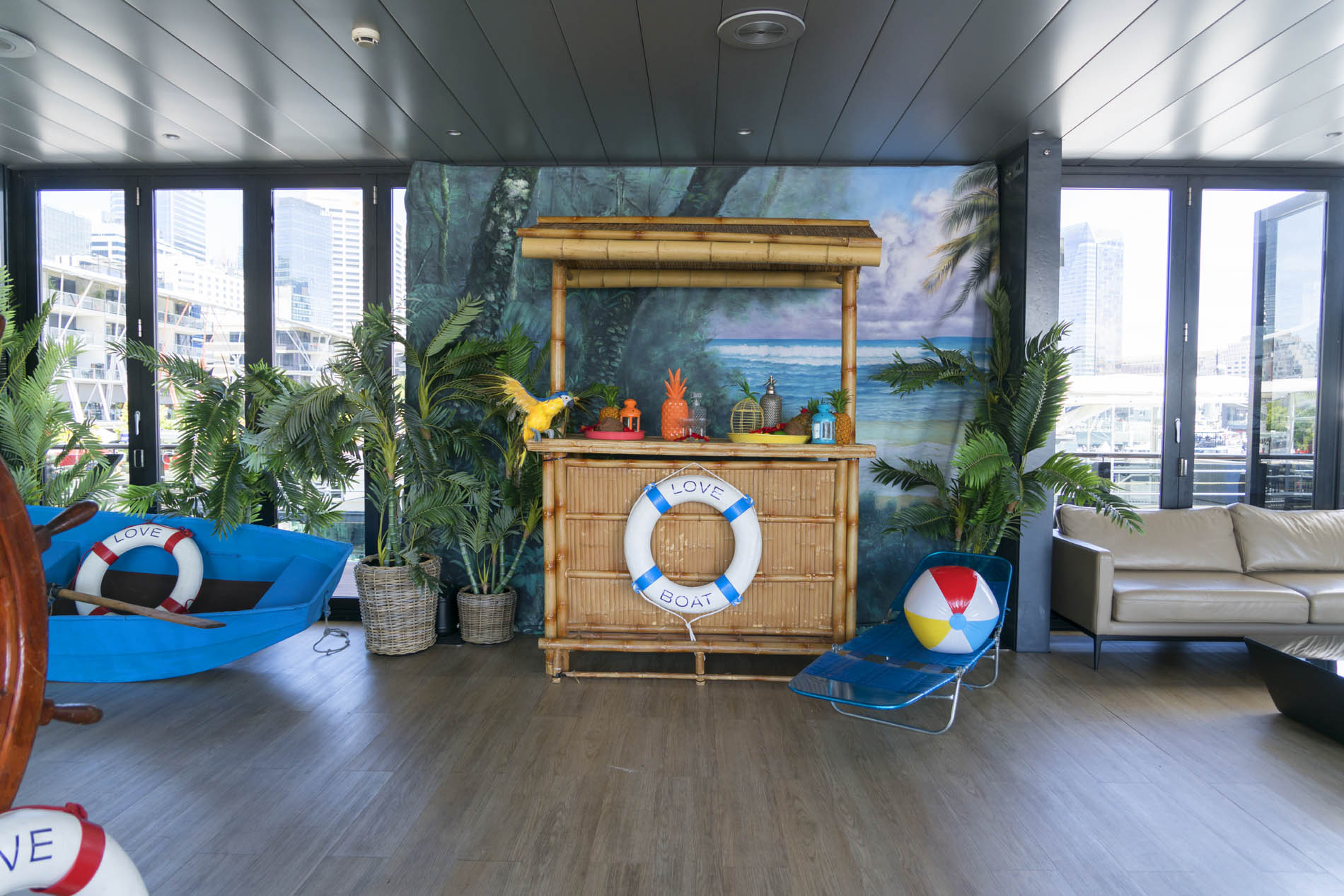 Bamboo bar with a life ring in a harbourside function room, with blue beanbags, palms and a beach ball