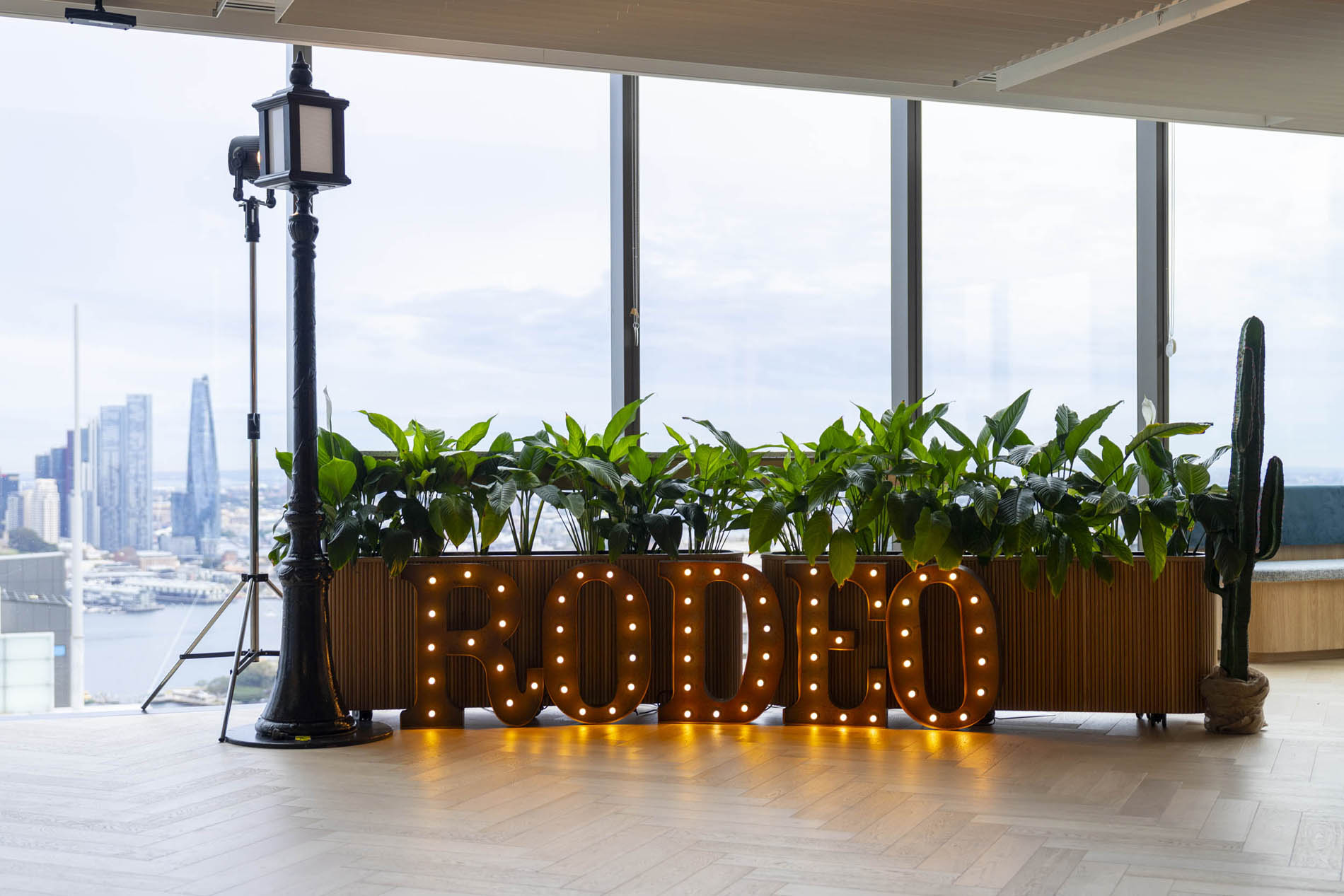 Illuminated marquee RODEO letters styled with potted greenery against a city skyline