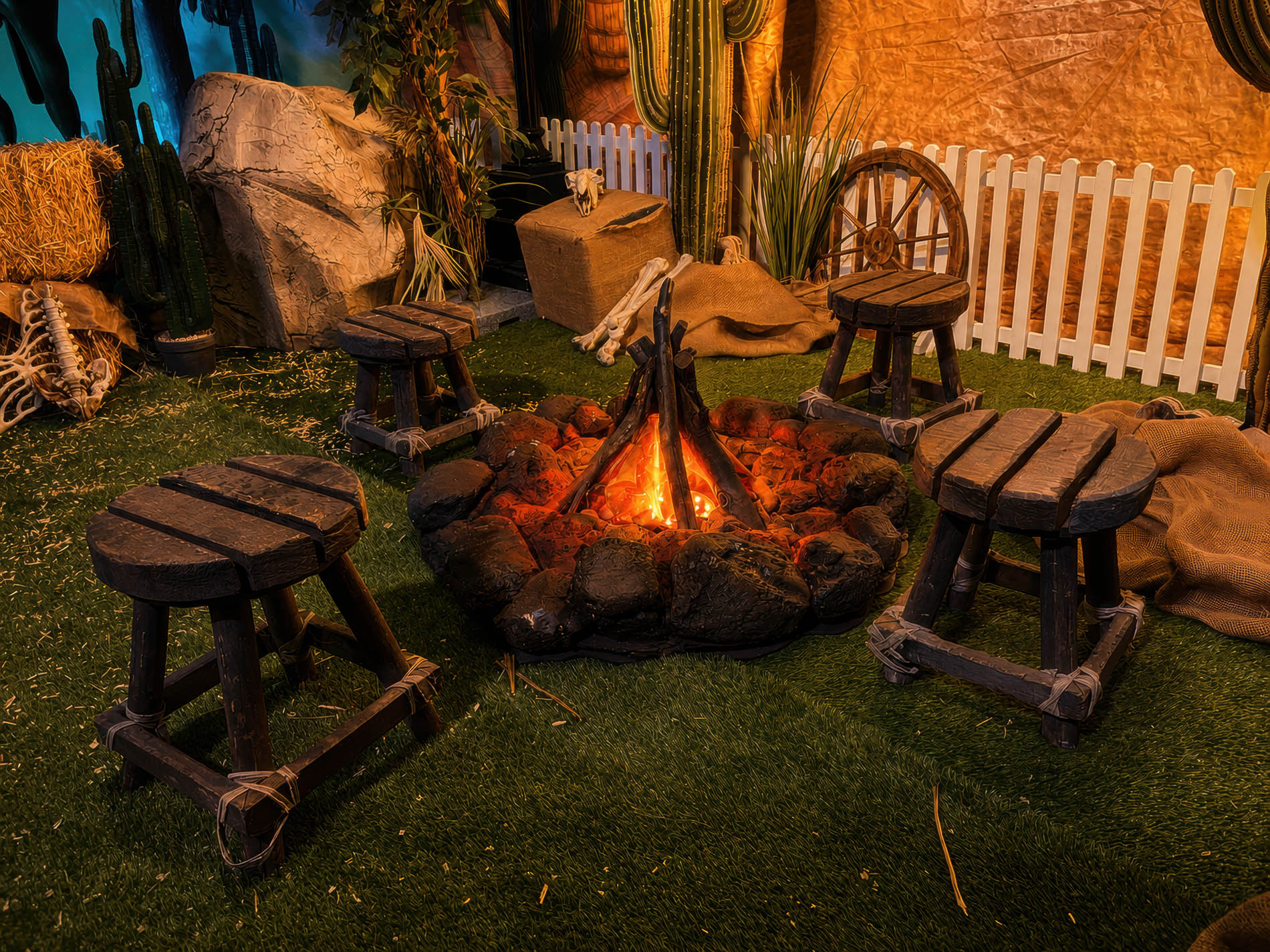 Glowing campfire ring with stump seating, hay bales, and saguaro cacti behind a white picket fence