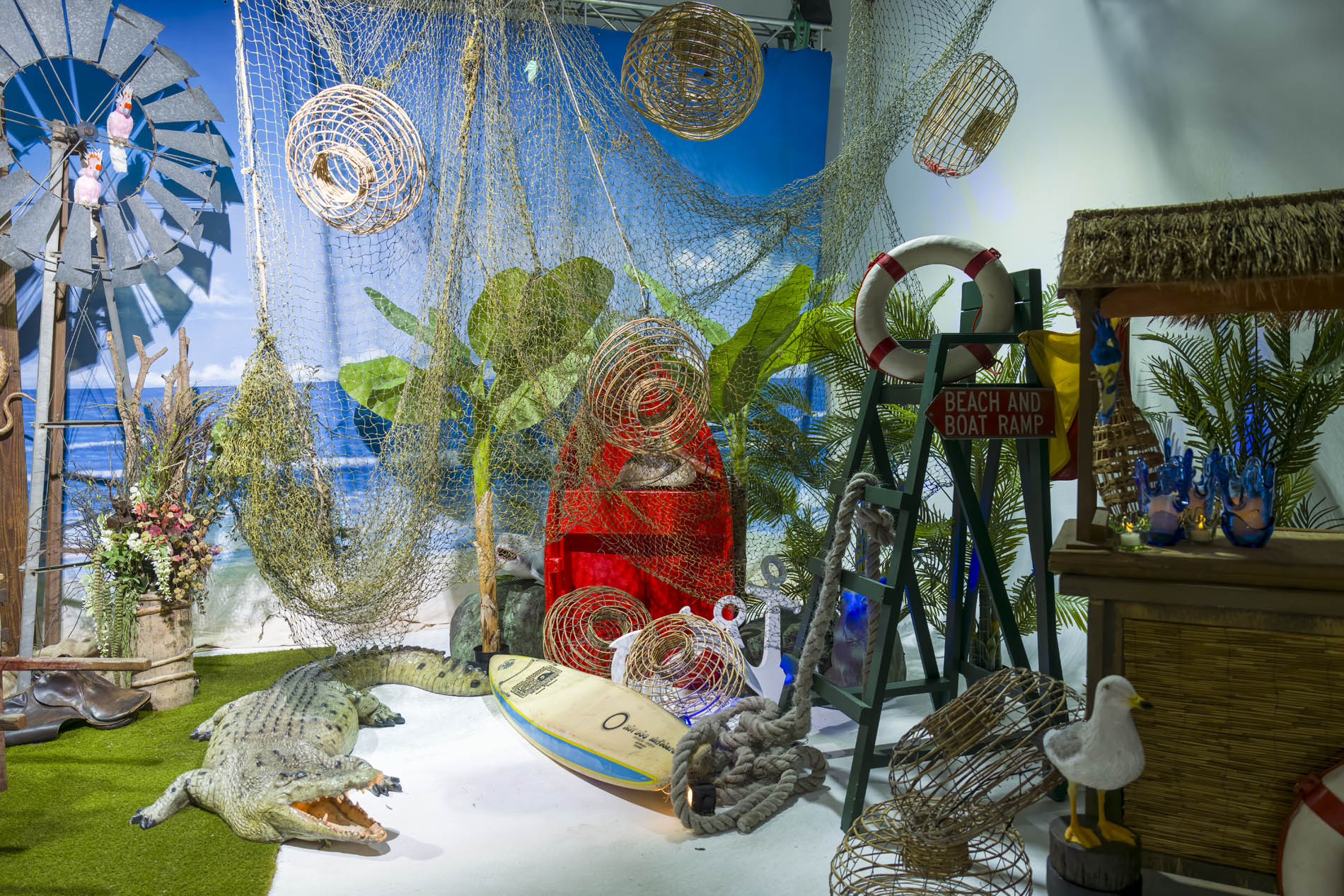 Boatshed scene with hanging fishing nets, wicker lobster pots, a red rowboat, windmill, seagulls and a crocodile on the sand