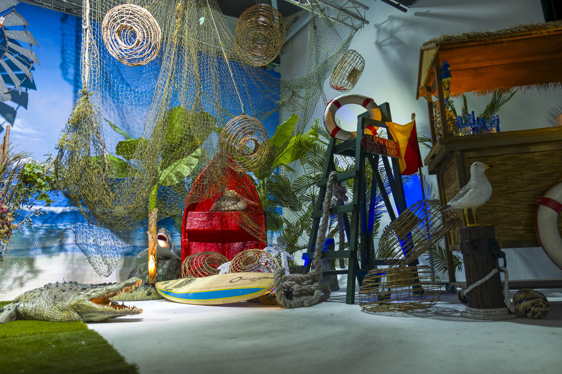 Wide boatshed scene with nets, lobster pots, a red rowboat, surfboard, crocodile and thatched shack bar